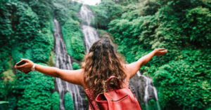 Young woman standing in front of a water fall with her arms outreached.