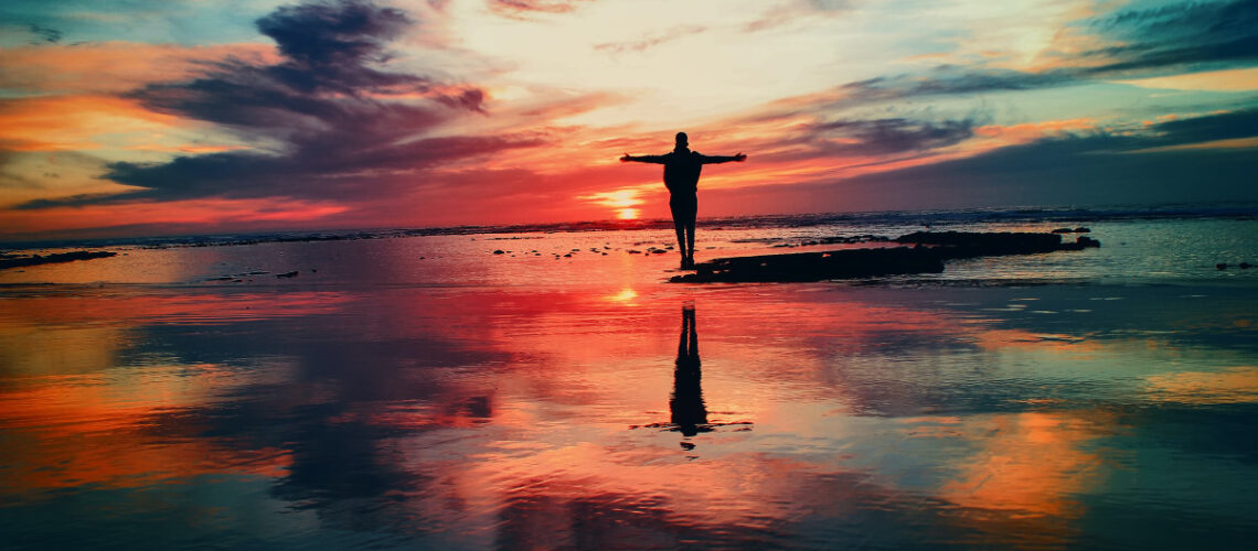 person with hands above their head on a beach watching a sunset