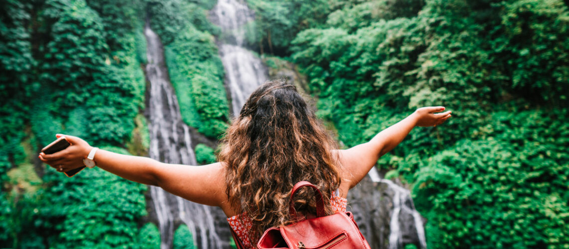 Young woman standing in front of a water fall with her arms outreached.