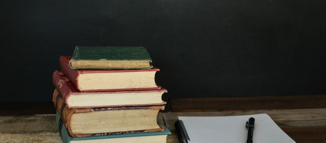 Books stacked on a table beside a pen on a notepad